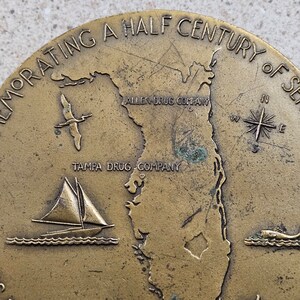 May include: A bronze commemorative medallion celebrating a half century of service. The medallion features a map of Florida with the text "Allen Drug Company" and "Tampa Drug Company" inscribed. A sailboat and a compass are also depicted.