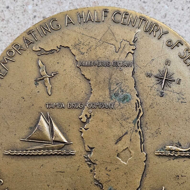 May include: A bronze commemorative medallion celebrating a half century of service. The medallion features a map of Florida with the text "Allen Drug Company" and "Tampa Drug Company" inscribed. A sailboat and a compass are also depicted.