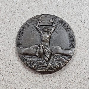 May include: A bronze commemorative medal with the text "IN MEMORY OF THE 6 MILLION VICTIMS." The medal features a central figure holding tablets, with figures below. The background includes clouds and a horizon line.