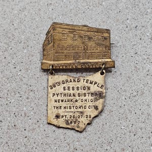 May include: Vintage gold-toned pin featuring a detailed building labeled "PYTHIAN CASTLE" above a pendant. The pendant reads "39TH GRAND TEMPLE SESSION PYTHIAN SISTERS NEWARK, OHIO THE HISTORIC CITY SEPT. 26-27-28 1927".