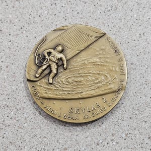 May include: A bronze medal commemorating the Skylab 2 mission. The medal features an astronaut in a spacesuit, a depiction of the Skylab space station, and the names of the crew members: Alan I Bean, Dr. Owen K. Garriott, and Jack R. Lousma.