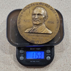 May include: A bronze commemorative medal with a portrait of a man in a suit and tie. The medal reads "Golden Anniversary * Tampa Drug Company * 1906 - 1956 * William George Allen 1865 - 1945 * Allen Drug Company" and is being weighed on a digital scale that reads 175.3 grams.