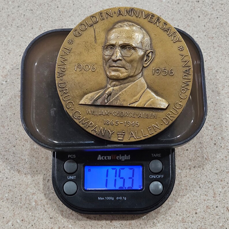 May include: A bronze commemorative medal with a portrait of a man in a suit and tie. The medal reads "Golden Anniversary * Tampa Drug Company * 1906 - 1956 * William George Allen 1865 - 1945 * Allen Drug Company" and is being weighed on a digital scale that reads 175.3 grams.