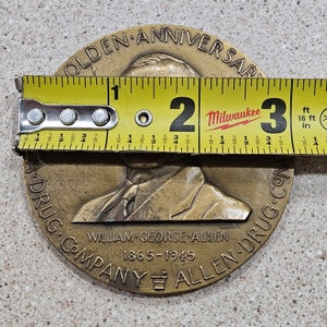 May include: A bronze commemorative coin celebrating the Golden Anniversary of William George Allen Drug Company. The coin features a portrait of a man in a suit and tie, with the text "Golden Anniversary" and "William George Allen Drug Company 1865-1945" around the edge.