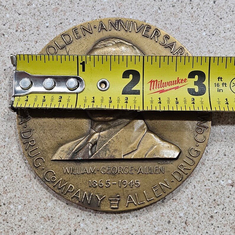 May include: A bronze commemorative coin celebrating the Golden Anniversary of William George Allen Drug Company. The coin features a portrait of a man in a suit and tie, with the text "Golden Anniversary" and "William George Allen Drug Company 1865-1945" around the edge.