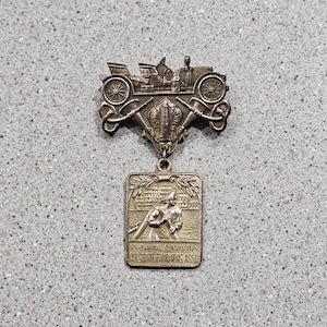 May include: Vintage silver-toned metal badge with a detailed design. The top features a classic car, and the bottom has a rectangular plaque with a figure and text. The text reads "55th Annual Convention NJ State Firemens Assn."