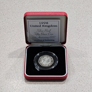May include: A red coin case with a silver proof fifty pence coin inside. The coin is from 1998 and commemorates the 25th anniversary of the EEC. The case has a label that reads "1998 United Kingdom Silver Proof Fifty Pence Coin 25th Anniversary EEC Certificate of Authenticity."