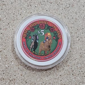 May include: A white plastic casino chip with a green background and red lettering. The chip features a picture of two horses and jockeys. The text on the chip reads "Fiesta Casino Hotel Las Vegas", "Derby of Champions", "May 6th, 2000", "Limited Edition 750", and "$5".