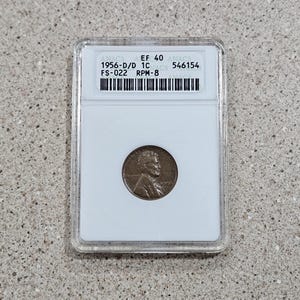 May include: A 1956-D/D 1C penny graded EF 40 by ANACS, encapsulated in a clear protective holder. The coin features a profile of a man and the text "1956". The holder has the text "ANACS" and a barcode.