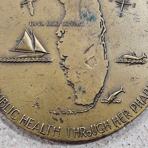 May include: A bronze coin with a map of Florida, a sailboat, a dolphin, and a fish. The text "TAMPA DRUG COMPANY" and "PUBLIC HEALTH THROUGH HER PHARMACY" are inscribed on the coin.