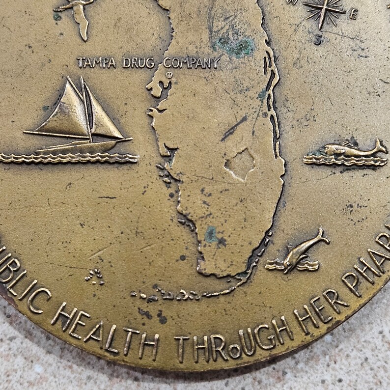 May include: A bronze coin with a map of Florida, a sailboat, a dolphin, and a fish. The text "TAMPA DRUG COMPANY" and "PUBLIC HEALTH THROUGH HER PHARMACY" are inscribed on the coin.