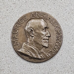 May include: A bronze-colored round medal featuring a profile portrait of a man with a beard. The text "MAURICE ANSIAUX" and "MCMXXXIX" are inscribed around the edge. The artist's signature, "dolf.Fecit 39", is at the bottom.