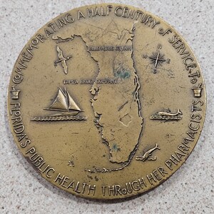 May include: A bronze commemorative medal celebrating a half century of service to Florida's public health through her pharmacists. The medal features a map of Florida, a sailboat, a compass, and a mortar and pestle.