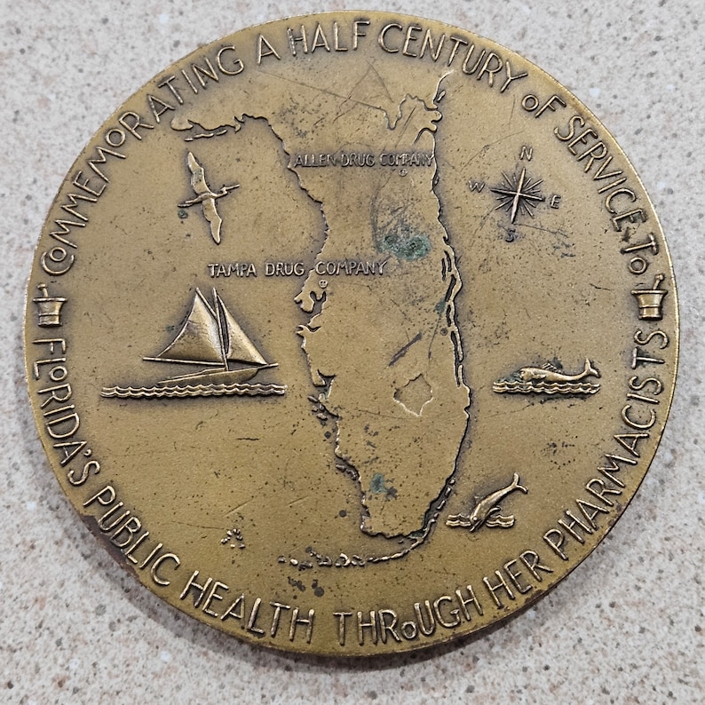 May include: A bronze commemorative medal celebrating a half century of service to Florida's public health through her pharmacists. The medal features a map of Florida, a sailboat, a compass, and a mortar and pestle.