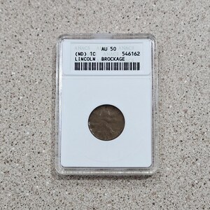 May include: A brown Lincoln cent coin encased in a clear plastic holder. The holder has a white insert with text that reads "ANACS AU 50" and "LINCOLN BROCKAGE". The coin is visible through a circular opening.