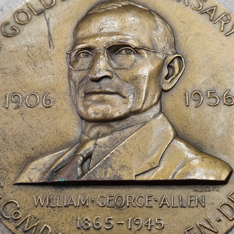 May include: A bronze medal with a portrait of a man wearing glasses. The medal has the text "1906" and "1956" on either side of the portrait. The text "WILLIAM GEORGE ALLEN 1865-1945" is at the bottom of the medal.