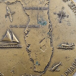May include: A bronze-colored metal token featuring a map of Florida with the text "Tampa Drug Company" and "Allen Drug Company" on the top. The token also features a compass, a sailboat, and two dolphins.