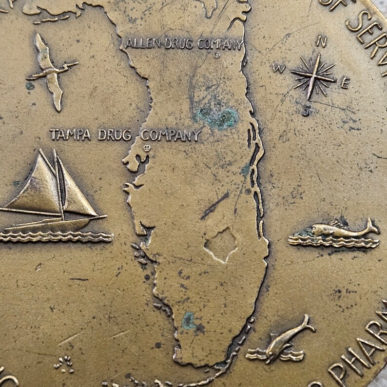 May include: A bronze-colored metal token featuring a map of Florida with the text "Tampa Drug Company" and "Allen Drug Company" on the top. The token also features a compass, a sailboat, and two dolphins.