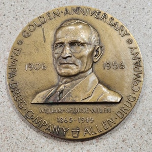 May include: A bronze medal commemorating the 50th anniversary of the Tampa Drug Company, featuring a portrait of William George Allen, the company's founder, and the text "Golden Anniversary 1906-1956 Tampa Drug Company Allen Drug Company William George Allen 1865-1945".