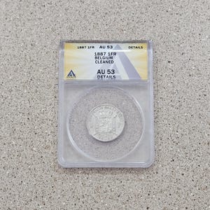 May include: A silver coin from 1887, encased in a clear plastic holder. The holder has a yellow and white label with text indicating the coin's details, including "1887 1FR BELGIUM CLEANED" and "AU 53 DETAILS". The coin is centered within the holder.