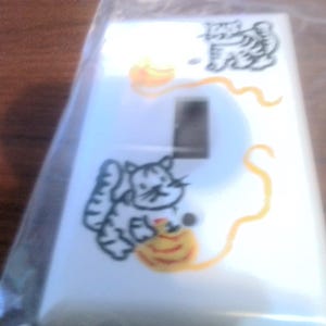 May include: White light switch cover with a hand-painted design of two cats playing with yarn. One cat is white and the other is orange.