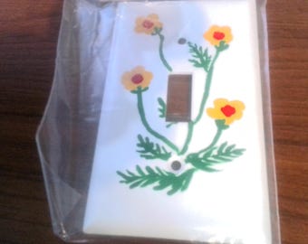 Puede incluir: Tapa de interruptor de luz blanca con un diseño pintado de flores amarillas con centros rojos y tallos verdes.