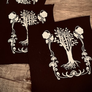 May include: Three black fabric patches with white embroidered designs of a stylized tree with flowers and mushrooms. The tree has a thick trunk and many branches with leaves. The flowers are simple and round. The mushrooms are stylized with a rounded top and a stem.