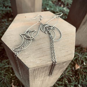 Può includere: Un paio di orecchini pendenti color argento con una forma a mezzaluna sospesa a un gancio. Ogni orecchino presenta una sottile cornice metallica curva e due delicate catenelle argentate che pendono verticalmente dal design.
