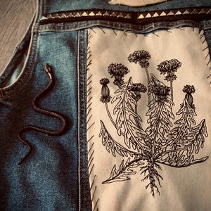 Pode incluir: Colete de ganga azul com um patch branco com uma ilustração de dente-de-leão a preto e branco. O colete é adornado com uma pequena cobra preta, um adorno em forma de osso e tachas prateadas.