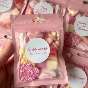 Peut inclure: Un sachet rose refermable rempli de bonbons gélifiés assortis, incluant des pièces en forme de cœur recouvertes de perles roses et blanches. Une étiquette ronde blanche porte l'inscription Bridesmaid avec une petite icône de cœur.