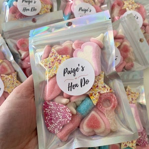 Peut inclure: Sacs transparents remplis de bonbons roses et blancs assortis, dont des c&oelig;urs, des &eacute;toiles et des anneaux. Chaque sac porte un autocollant circulaire avec l'inscription "Paige's Hen Do". Les bonbons sont de textures et de formes vari&eacute;es.