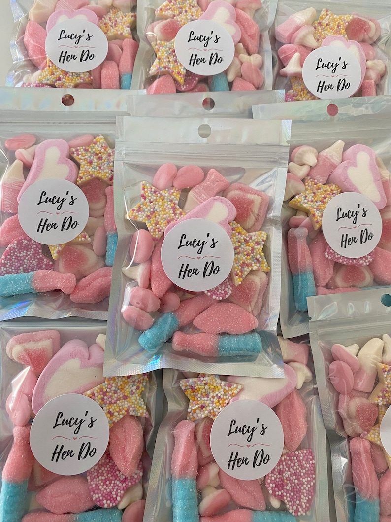 Peut inclure: Plusieurs sachets transparents remplis de bonbons roses, blancs et bleus. Chaque sachet porte un autocollant rond avec l'inscription "Lucy's Hen Do". Les bonbons comprennent des &eacute;toiles saupoudr&eacute;es et des guimauves.