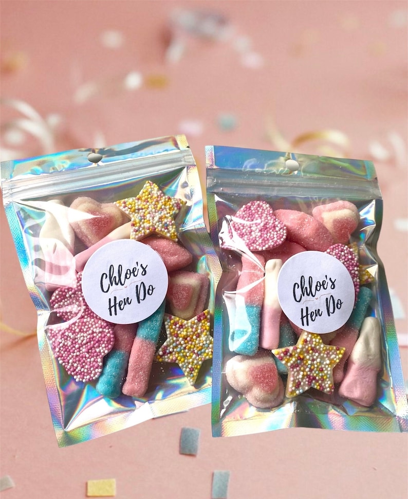 Peut inclure: Deux sachets transparents remplis de bonbons assortis, dont des formes d'&eacute;toiles et de c&oelig;urs, et de longs morceaux cylindriques. Chaque sachet porte un autocollant rond blanc avec l'inscription "Chloe's Hen Do". Les sachets ont un aspect holographique.