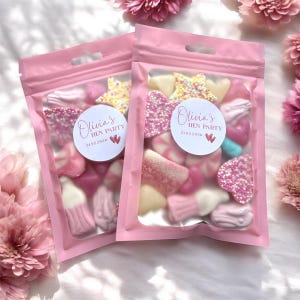 Peut inclure: Deux sachets roses en cellophane remplis de bonbons assortis. Chaque sachet porte un autocollant rond avec l'inscription "Olivia's HEN PARTY" et la date "21.02.2020". Les bonbons sont roses, blancs et jaunes, en forme d'étoiles et de cœurs.