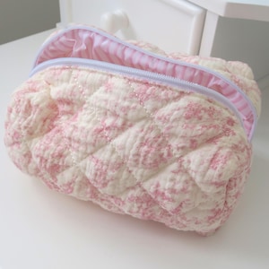 Könnte beinhalten: Eine gesteppte Kosmetiktasche aus weichem, strukturiertem Stoff. Die Tasche hat ein Muster aus rosa Blumenformen auf cremefarbenem Hintergrund. Das Innenfutter ist rosa und weiß gestreift. Die Tasche ist geöffnet und gibt den Blick ins Innere frei.