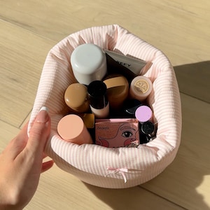 Peut inclure: Un panier en tissu à rayures roses et blanches rempli de divers produits cosmétiques. Le panier contient des contenants de différentes formes et tailles, notamment des pots, des tubes et des poudriers. Le mot "benefit" est visible sur un des poudriers.