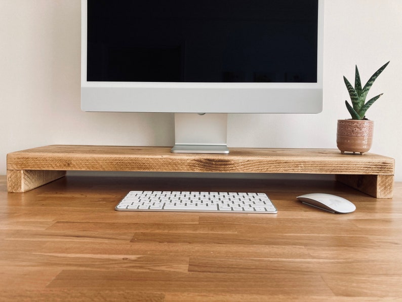 Pu&ograve; includere: Un supporto per monitor in legno con una tastiera bianca e un mouse bianco su una scrivania in legno. Una pianta grassa in vaso &egrave; sulla scrivania dietro il supporto.