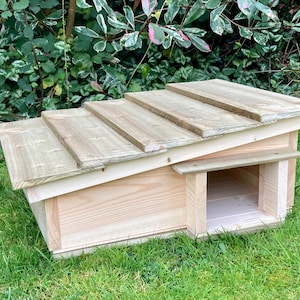 May include: A wooden hedgehog house with a slanted roof. The house is constructed from light-coloured wood and features a rectangular entrance. It is positioned on a grassy lawn, with green foliage in the background. The roof is made of overlapping wooden planks.