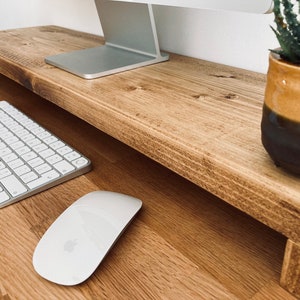 Pu&ograve; includere: Un supporto per monitor in legno con una tastiera e un mouse bianchi su una scrivania in legno. Una pianta grassa in vaso si trova sul lato destro del supporto.
