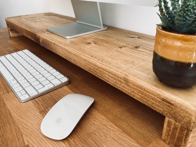 Pu&ograve; includere: Un supporto per monitor in legno con una tastiera e un mouse bianchi su una scrivania in legno. Una pianta grassa in vaso si trova sul lato destro del supporto.