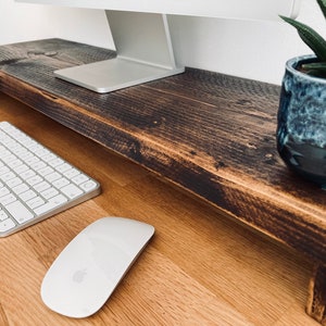 Pu&ograve; includere: Un supporto per monitor in legno marrone scuro con una tastiera e un mouse bianchi su una scrivania in legno chiaro. Una piccola pianta grassa in vaso &egrave; sul supporto.