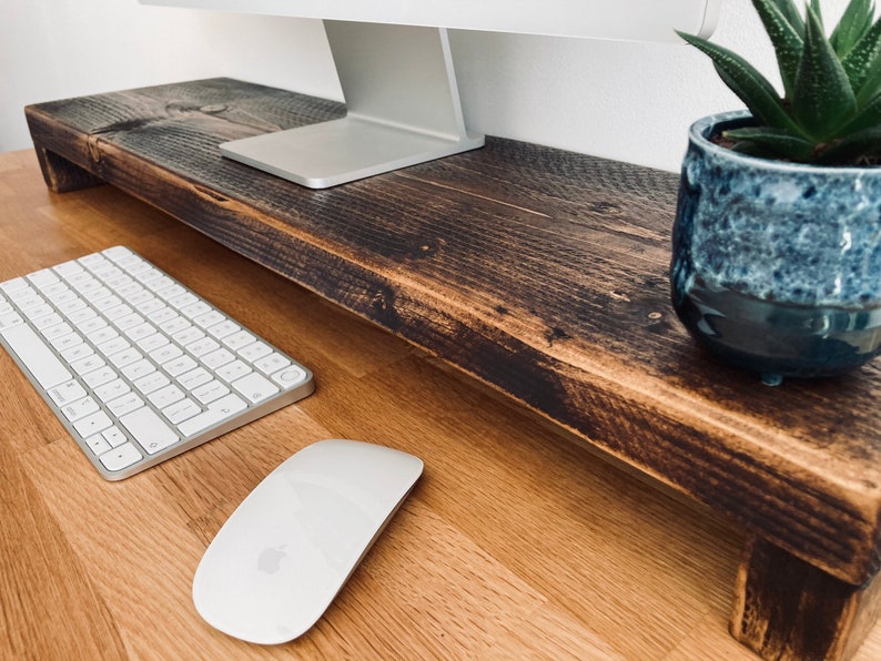 Pu&ograve; includere: Un supporto per monitor in legno marrone scuro con una tastiera e un mouse bianchi su una scrivania in legno chiaro. Una piccola pianta grassa in vaso &egrave; sul supporto.