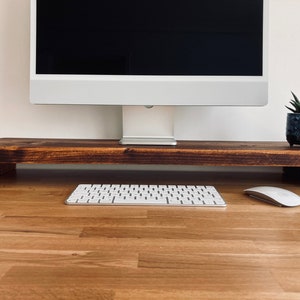 Pu&ograve; includere: Un supporto per monitor in legno con una tinta marrone scuro. Il supporto &egrave; posto su una scrivania in legno chiaro con una tastiera bianca e un mouse bianco. Una piccola pianta in vaso si trova a destra del supporto.