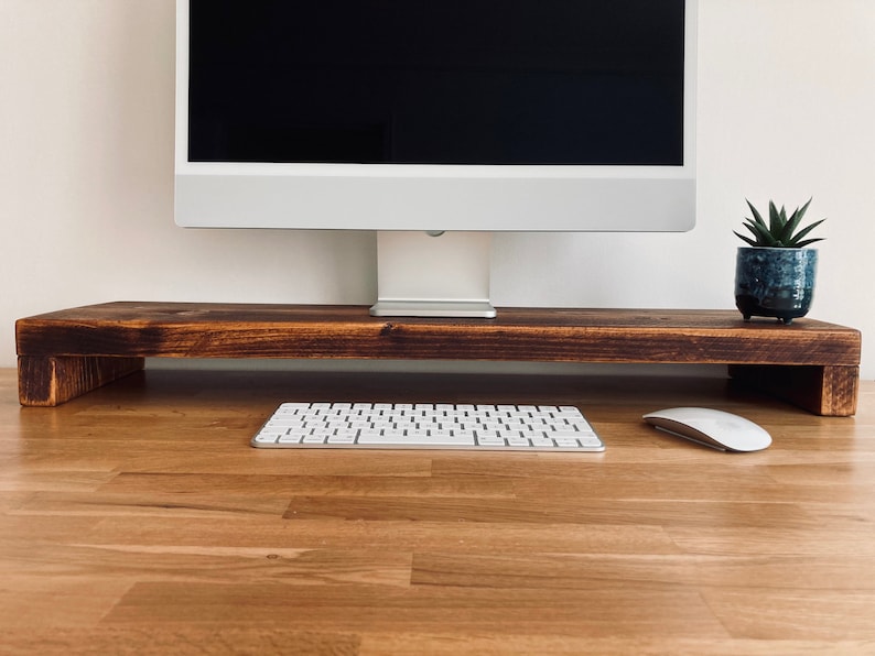 Pu&ograve; includere: Un supporto per monitor in legno con una tinta marrone scuro. Il supporto &egrave; posto su una scrivania in legno chiaro con una tastiera bianca e un mouse bianco. Una piccola pianta in vaso si trova a destra del supporto.