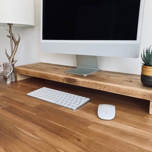 May include: A wooden monitor stand with a rustic finish, featuring a computer monitor, keyboard, and mouse. A small potted succulent plant sits on the right side of the stand.