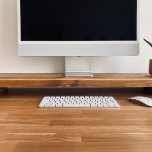 Pu&ograve; includere: Un supporto per monitor in legno con una finitura marrone chiaro. Il supporto &egrave; rialzato rispetto alla superficie e ha una forma rettangolare. Una tastiera bianca e un mouse bianco sono sulla superficie sotto il supporto.