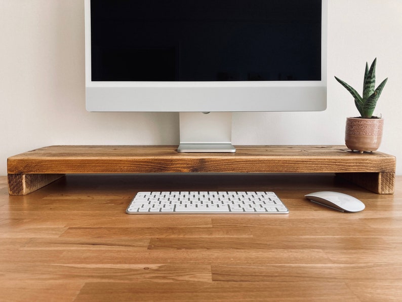 Pu&ograve; includere: Un supporto per monitor in legno con una finitura marrone chiaro. Il supporto &egrave; rialzato rispetto alla superficie e ha una forma rettangolare. Una tastiera bianca e un mouse bianco sono sulla superficie sotto il supporto.
