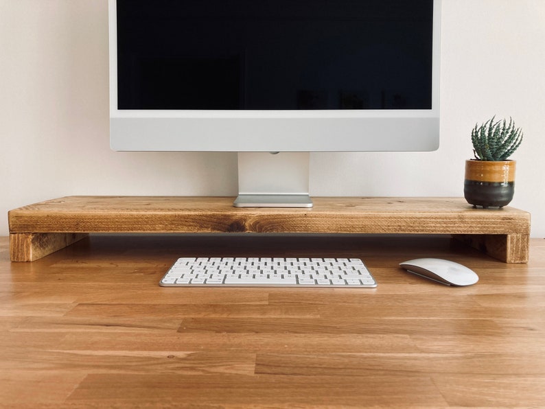 Pu&ograve; includere: Un supporto per monitor in legno con una finitura marrone chiaro. Una tastiera bianca e un mouse bianco sono sulla scrivania davanti al supporto. Una piccola pianta grassa in vaso si trova sul lato destro del supporto.