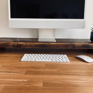 Pu&ograve; includere: Un supporto per monitor in legno marrone scuro con una tastiera bianca e un mouse bianco su una scrivania in legno marrone chiaro.