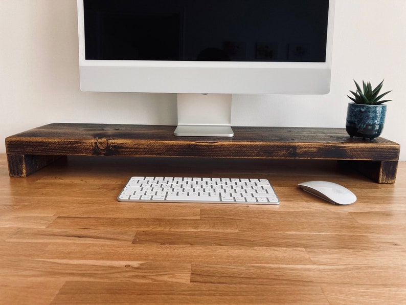 Pu&ograve; includere: Un supporto per monitor in legno marrone scuro con una tastiera bianca e un mouse bianco su una scrivania in legno marrone chiaro.